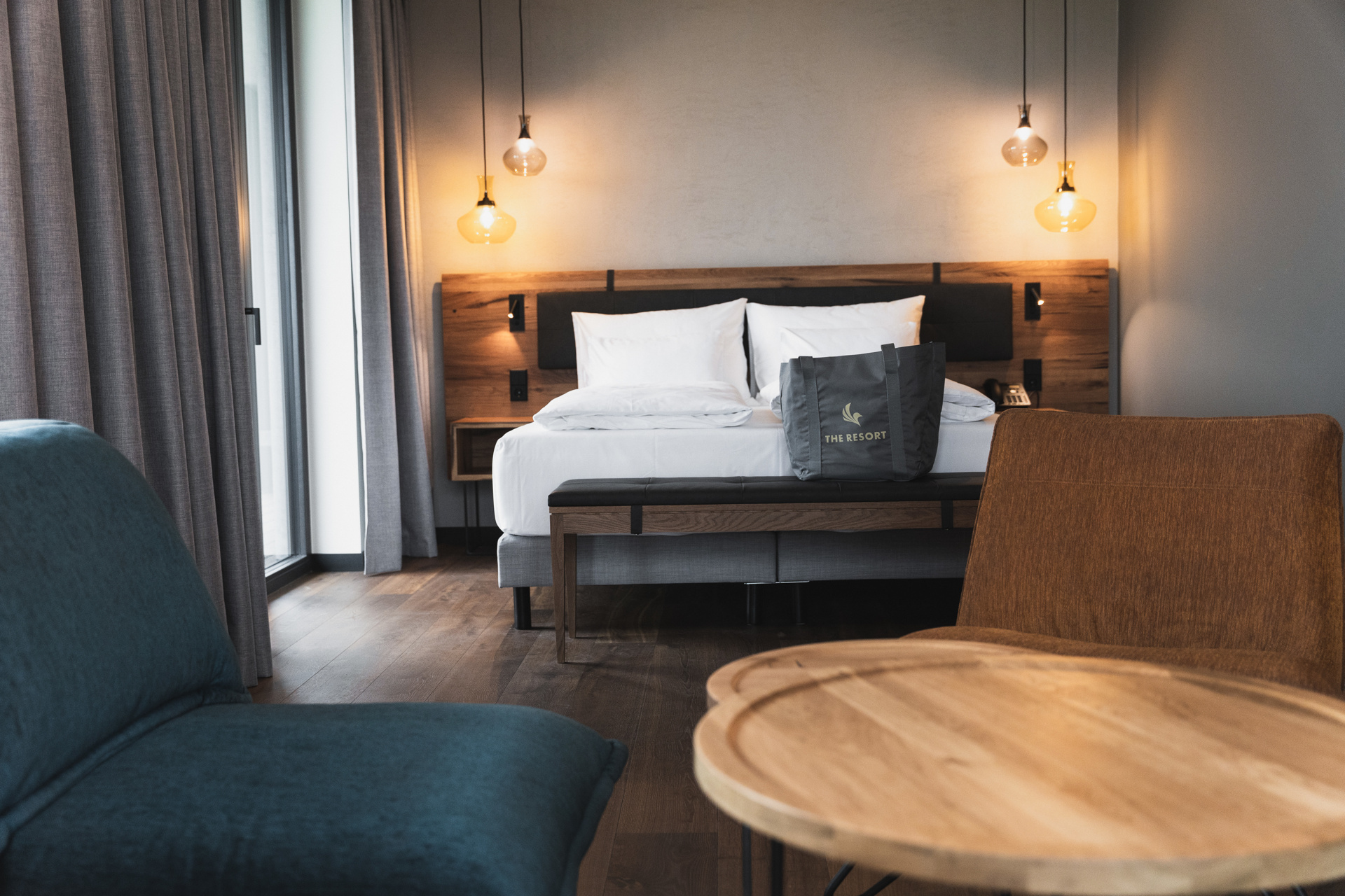 Ein modernes Hotelzimmer mit einem großen Bett, weißem Bettzeug, einem hölzernen Kopfteil, hängenden Pendelleuchten, einem grauen Sessel, einem Holztisch und einer Tragetasche auf dem Bett. Licht strömt durch ein Fenster mit grauen Vorhängen.
