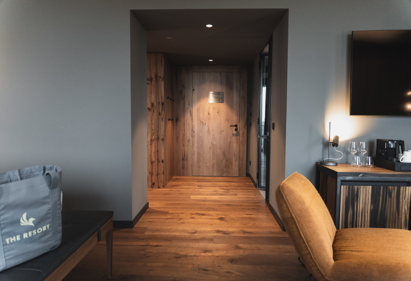 Moderner Hotelzimmereingang mit Holzboden und -tür, braunem Stuhl, Fernseher und einer grauen Tasche mit der Aufschrift The Resort.