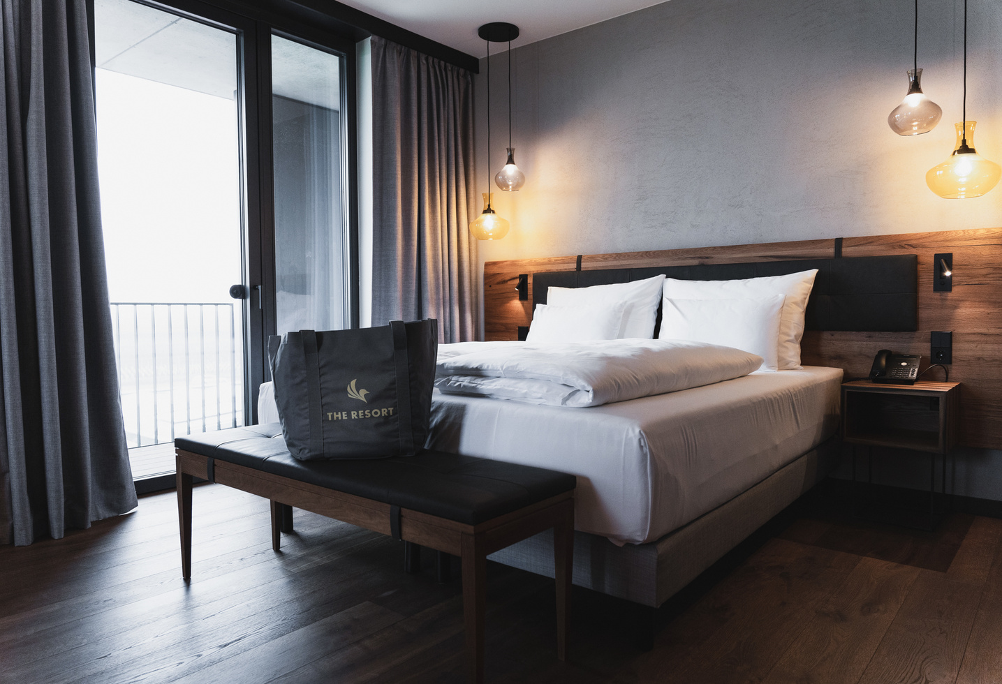 Modernes Hotelzimmer mit einem großen Bett, Hängelampen und einer Tragetasche mit der Aufschrift The Resort auf einer Bank.