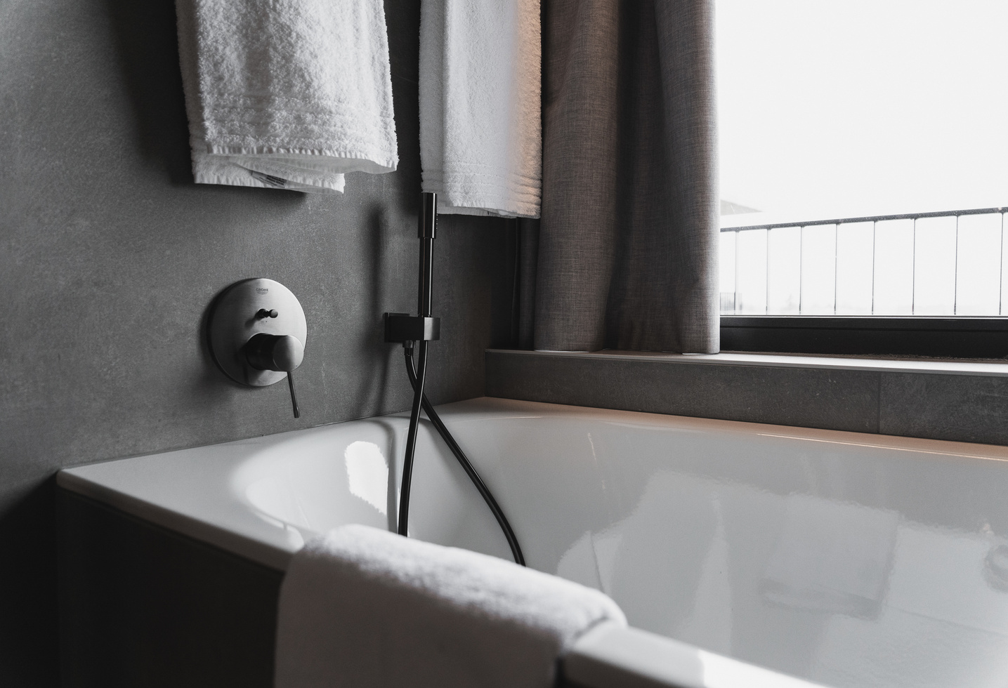 Moderne Badewanne mit Handbrause, weiße Handtücher und ein großes Fenster mit grauen Vorhängen.