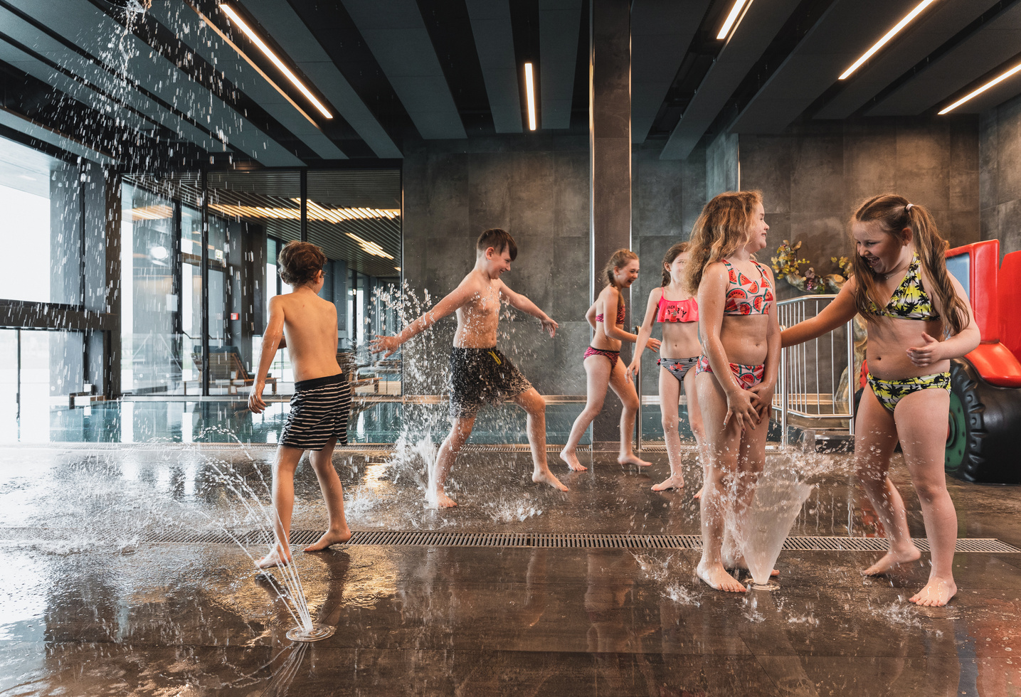 Kinder in Badeanzügen spielen und lachen gemeinsam in einem überdachten Wasserbereich mit Springbrunnen, die Wasser versprühen.