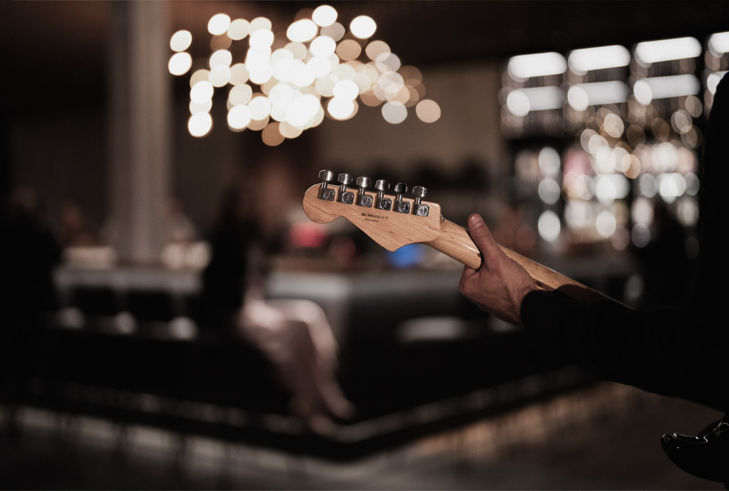 Eine Person hält eine E-Gitarre in einer schwach beleuchteten Bar mit verschwommenen Lichtern und Menschen im Hintergrund.