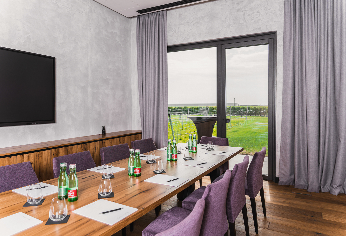 Ein moderner Konferenzraum mit einem Holztisch, lilafarbenen Stühlen und einem Blick auf grüne Felder durch große Fenster.
