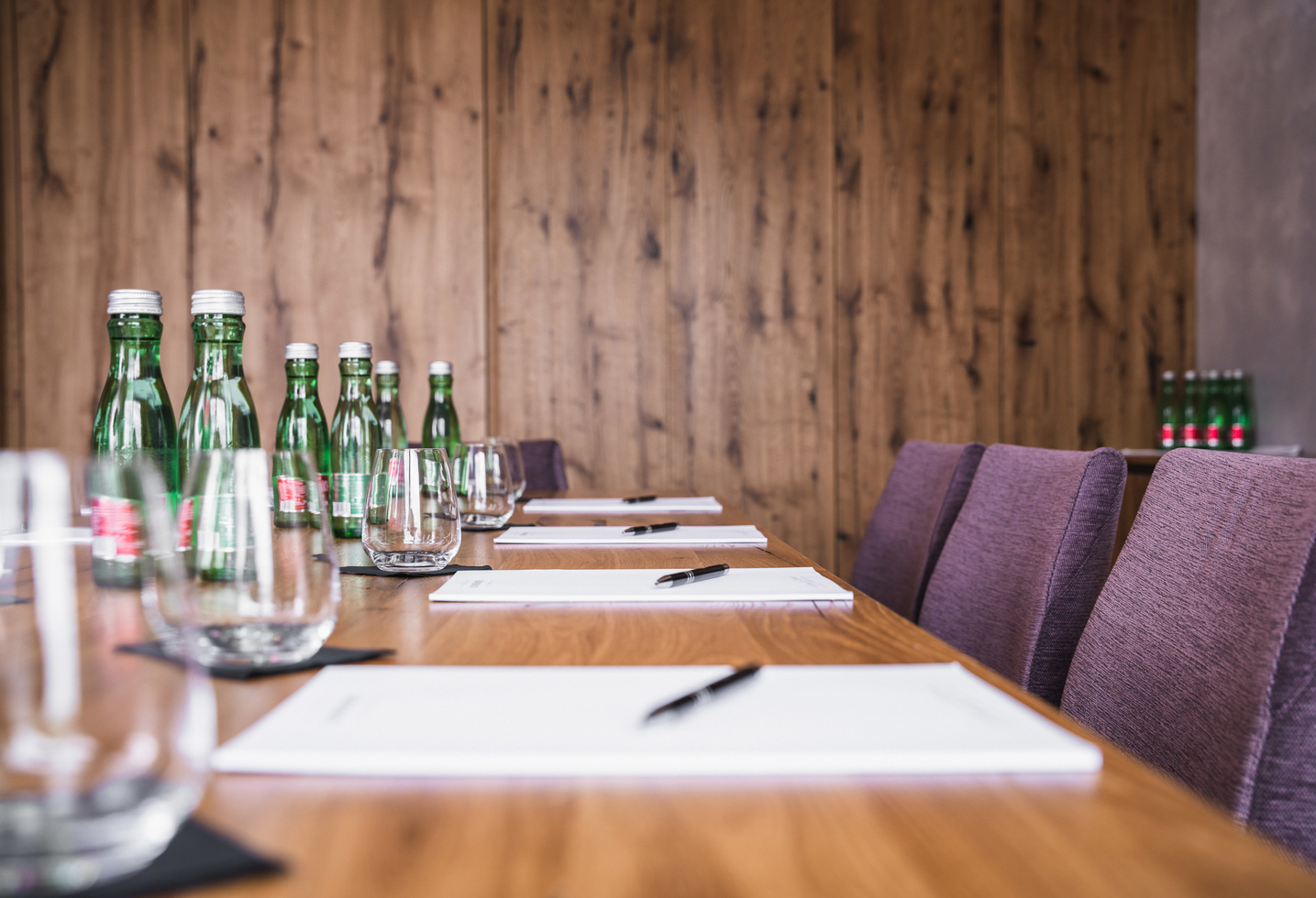 Nahaufnahme eines Konferenztisches mit Wasserflaschen, Gläsern, Notizbüchern und Stiften; lila Stühle am Tisch.