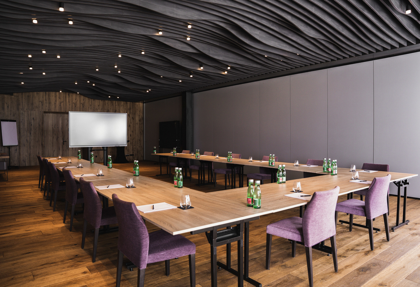 Moderner Konferenzraum mit U-förmigen Tischen, lilafarbenen Stühlen, Notizblöcken, Wasserflaschen und einer Projektionsfläche.