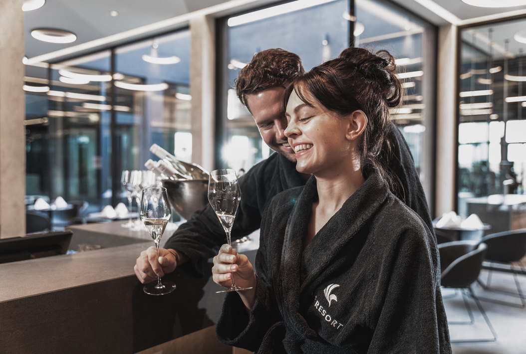 Ein lächelndes Paar in Bademänteln genießt Champagner in einer modernen Resort-Lounge und hält die Gläser zusammen.