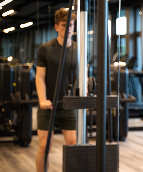 Eine Person benutzt eine Gewichtsmaschine in einem Fitnessstudio, im Hintergrund sind Fitnessgeräte zu sehen.