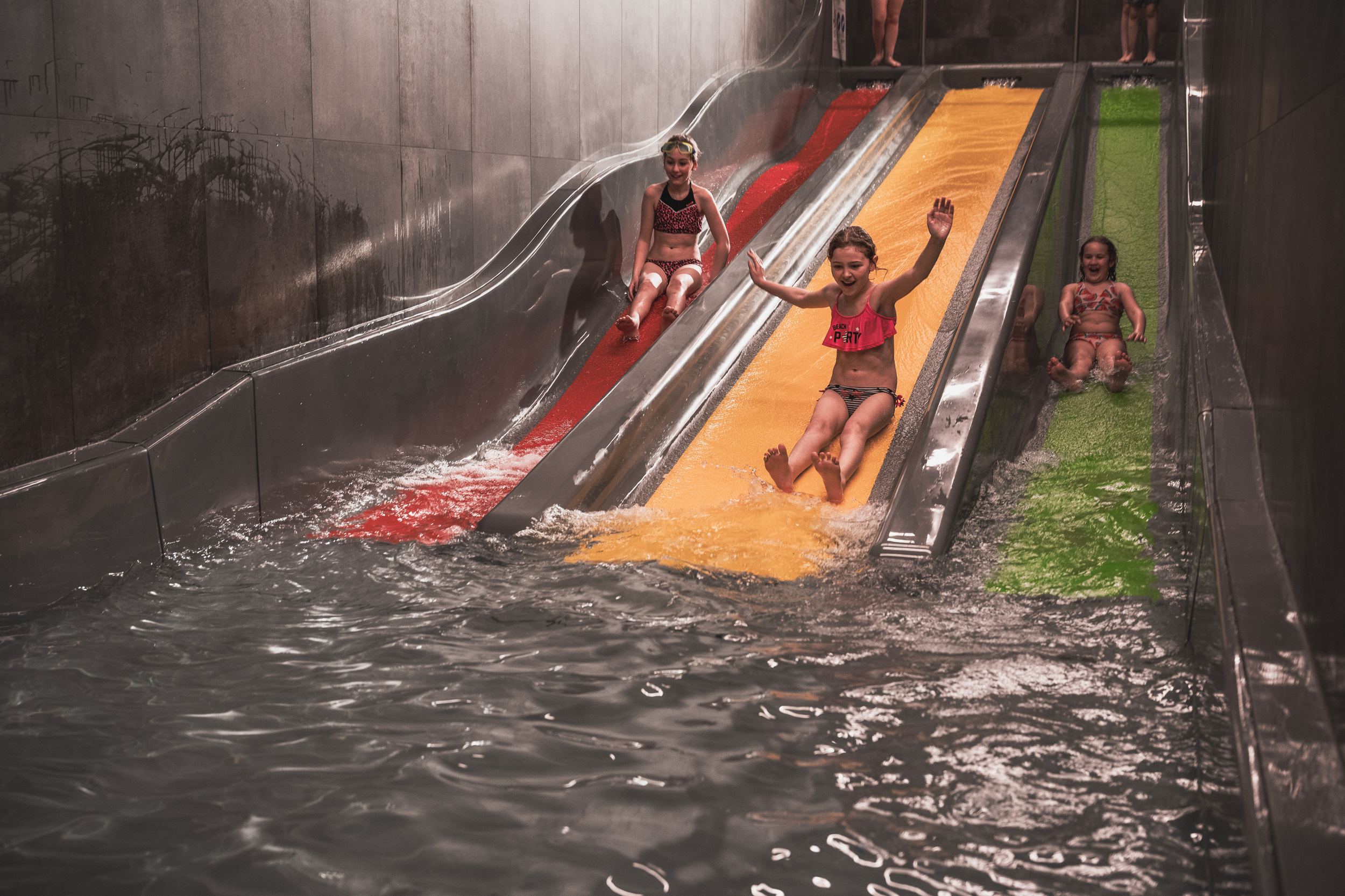 Vier Kinder in Badeanzügen rutschen über bunte Wasserrutschen in ein Becken in einem Indoor-Wasserpark.