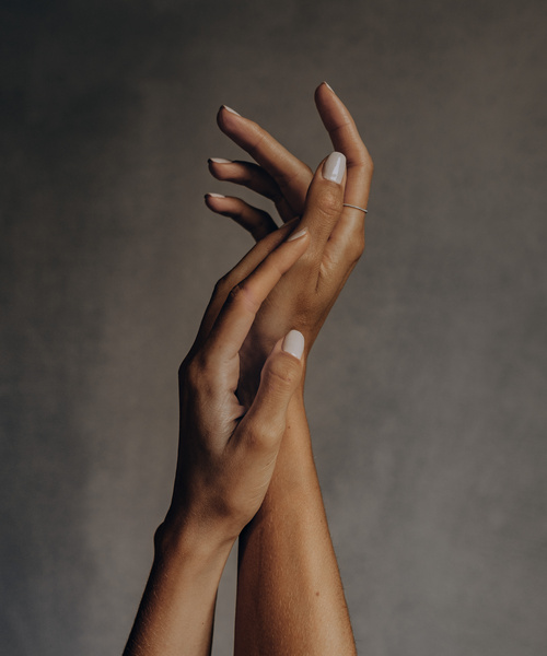 Zwei Hände mit hellem Nagellack berühren sich sanft vor einem schlichten, neutralen Hintergrund.