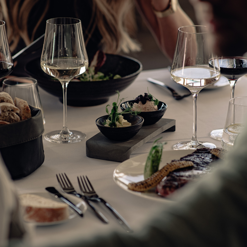 A table set with wine glasses, salad, bread, steak and small dishes, where people eat together.
