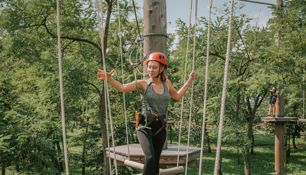 Woman with helmet crossing a rope bridge under trees in an outdoor adventure park.