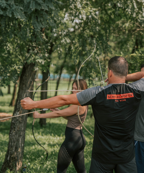 Drei Personen üben sich in einem Waldgebiet im Bogenschießen und zielen mit ihren Bögen auf unsichtbare Ziele.