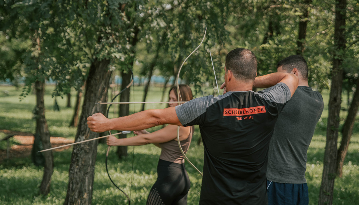 Three people are practicing archery in a wooded area, aiming their bows at invisible targets.