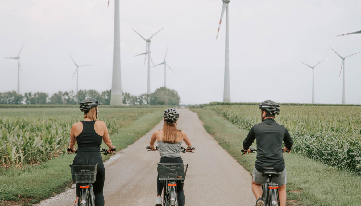 Three people cycle along a rural path lined with wind turbines and green fields under a cloudy sky.