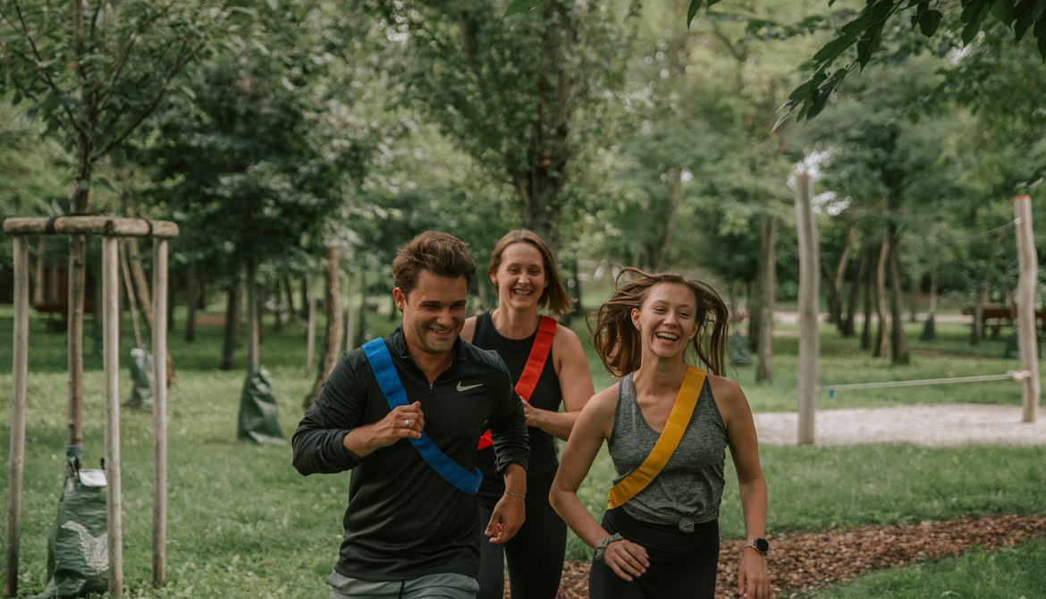 Three smiling adults jogging in a green park, each wearing a colored sash across their chest.