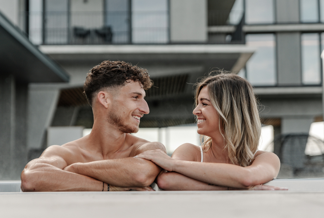 Lächelnder Mann und lächelnde Frau in einem Pool, die sich an den Rand lehnen und sich liebevoll ansehen.