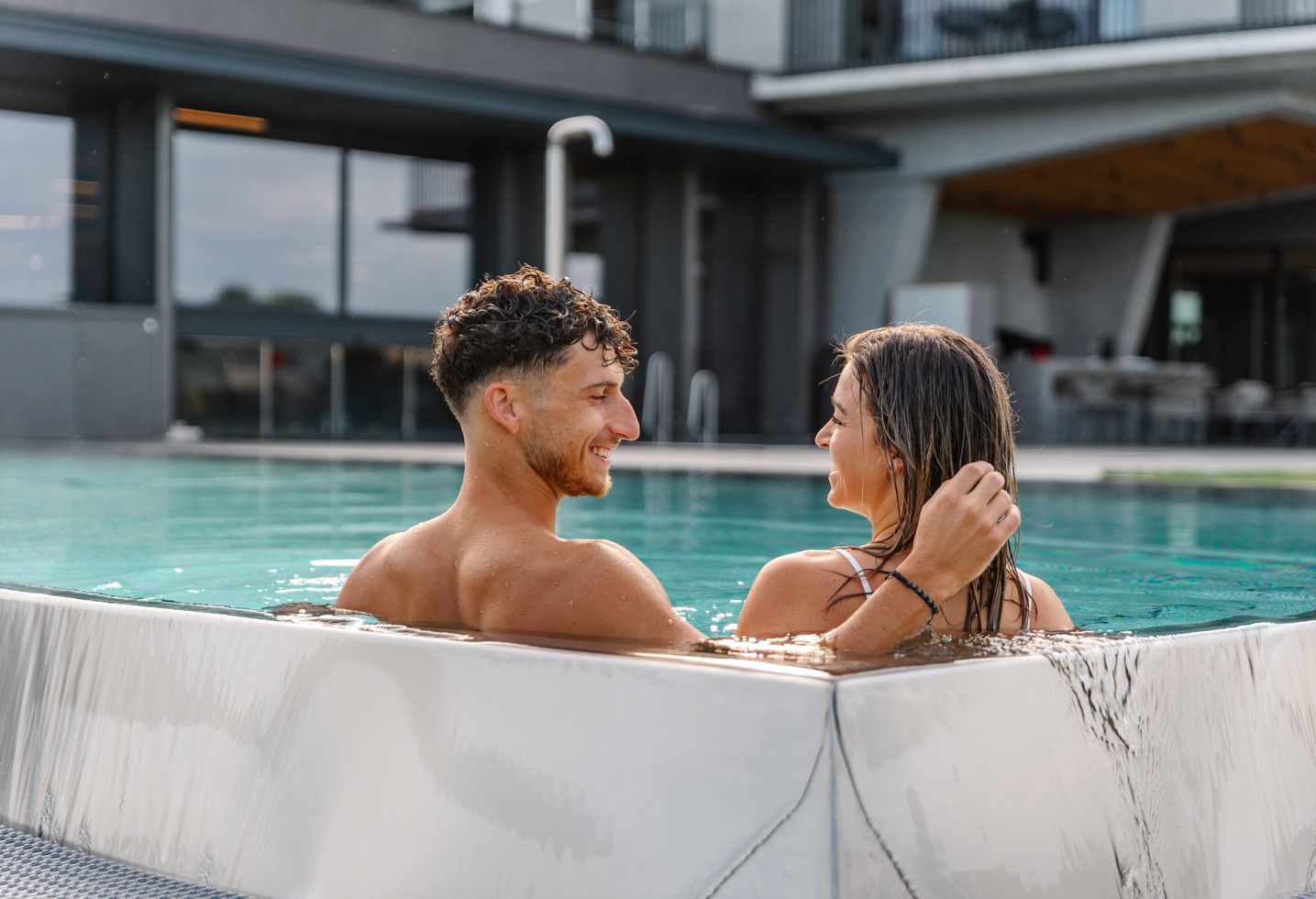 Ein lächelndes Paar entspannt sich gemeinsam in einem Außenpool, während es sich gegenübersteht und den Moment genießt.