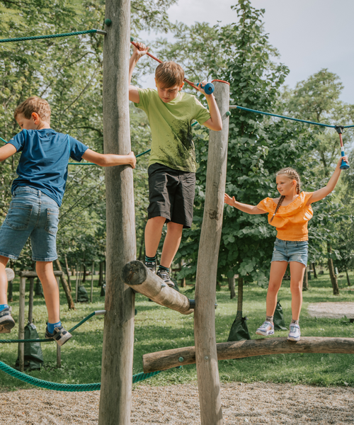 Drei Kinder balancieren und klettern auf Holzkonstruktionen auf einem von Bäumen umgebenen Spielplatz im Freien.