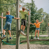Drei Kinder balancieren und klettern auf Holzkonstruktionen auf einem von Bäumen umgebenen Spielplatz im Freien.