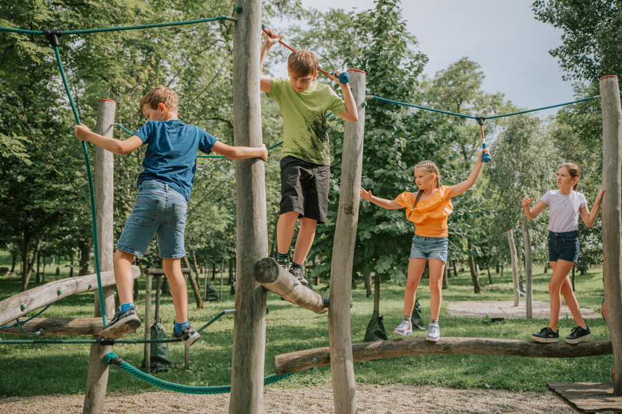 Three children balance and climb on wooden structures in an outdoor playground surrounded by trees.