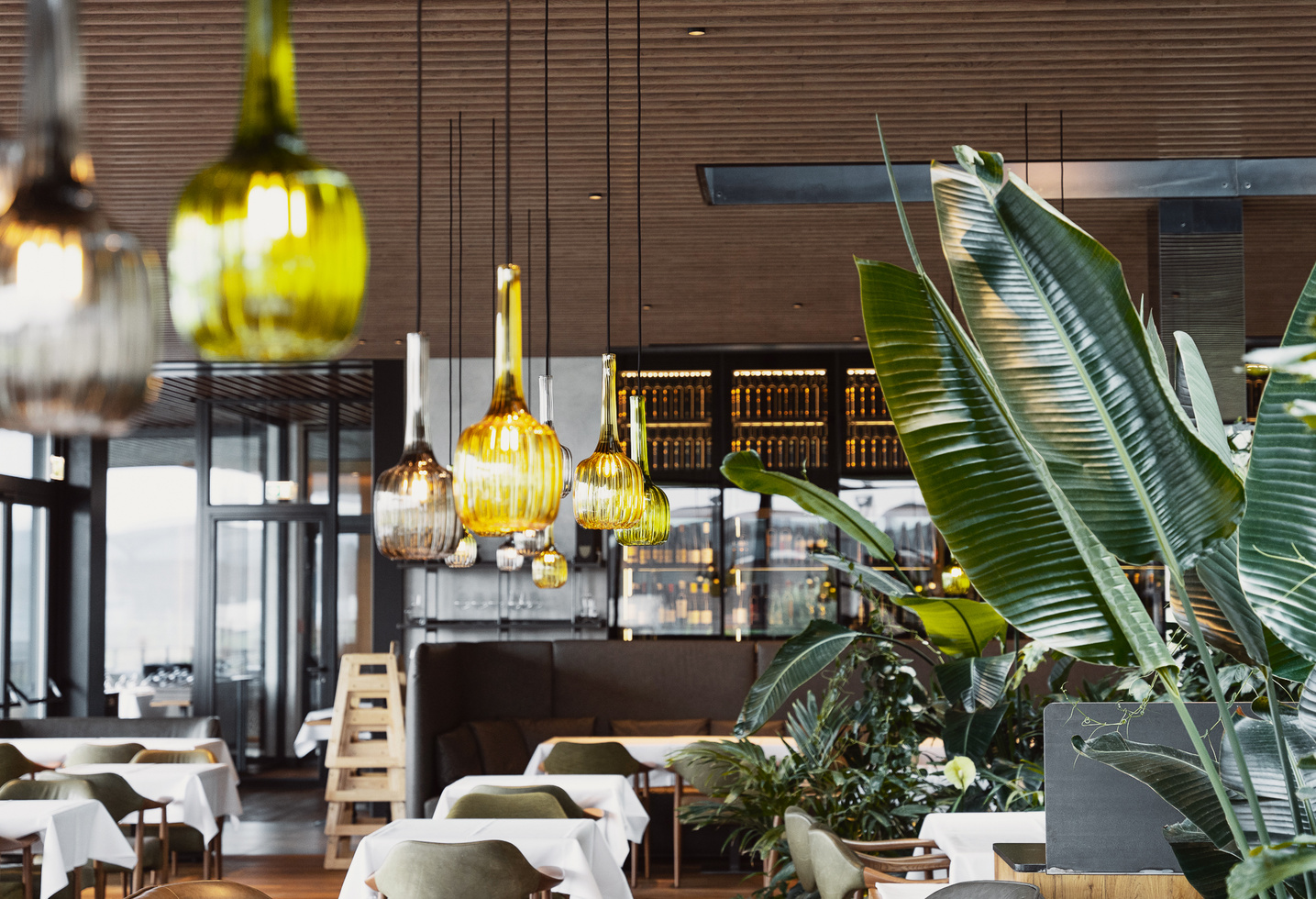 Modern restaurant interior with green plants, yellow pendant lights and tables with white tablecloths.