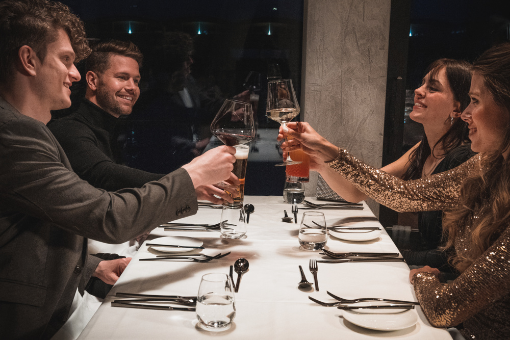 Vier Personen sitzen an einem Tisch, stoßen mit Gläsern an und lächeln, in einem warm beleuchteten, eleganten Ambiente.