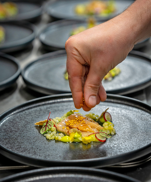 A hand sprinkles spices on a gourmet dish with vegetables and garnishes on a black plate.
