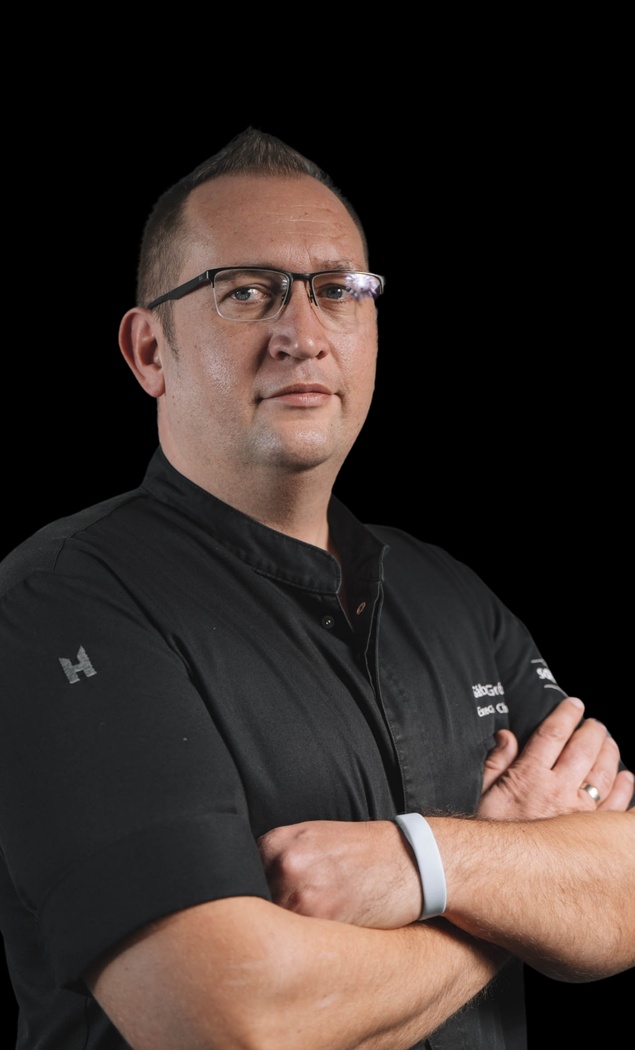 A chef in a black uniform and glasses stands with his arms folded in front of a black background.
