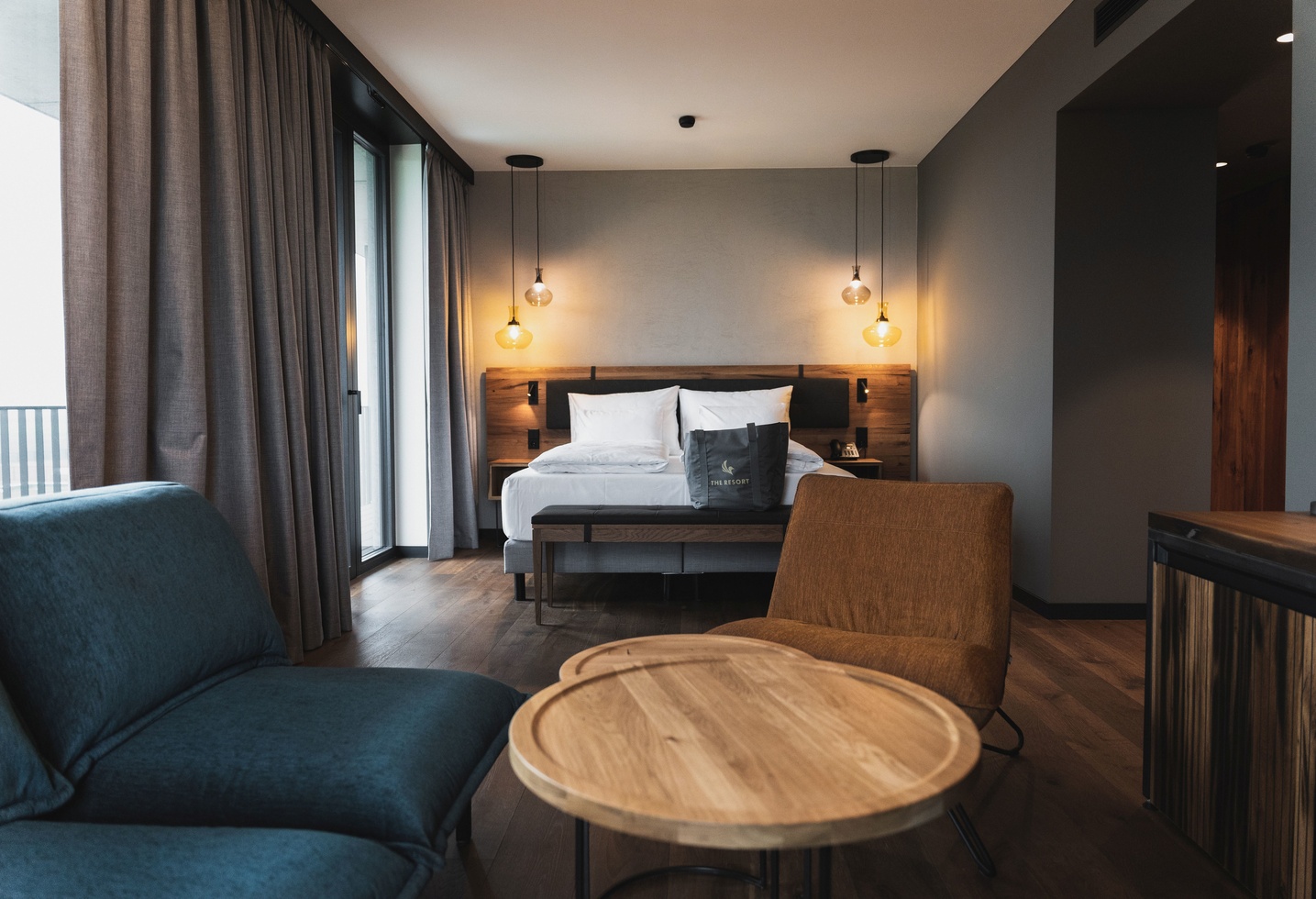 Modernes Hotelzimmer mit einem Doppelbett, runden Holztischen, blauem Sofa und warmer Beleuchtung.