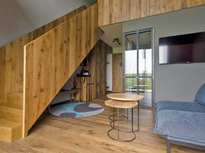Moderner Wohnraum mit Holztreppe und -fußboden, einem blauen Sofa, zwei runden, ineinander verschachtelten Tischen, einem an der Wand montierten Fernseher und einer Glastür, die auf einen Balkon führt. Ein bunter Teppich setzt Akzente in der gemütlichen Einrichtung.