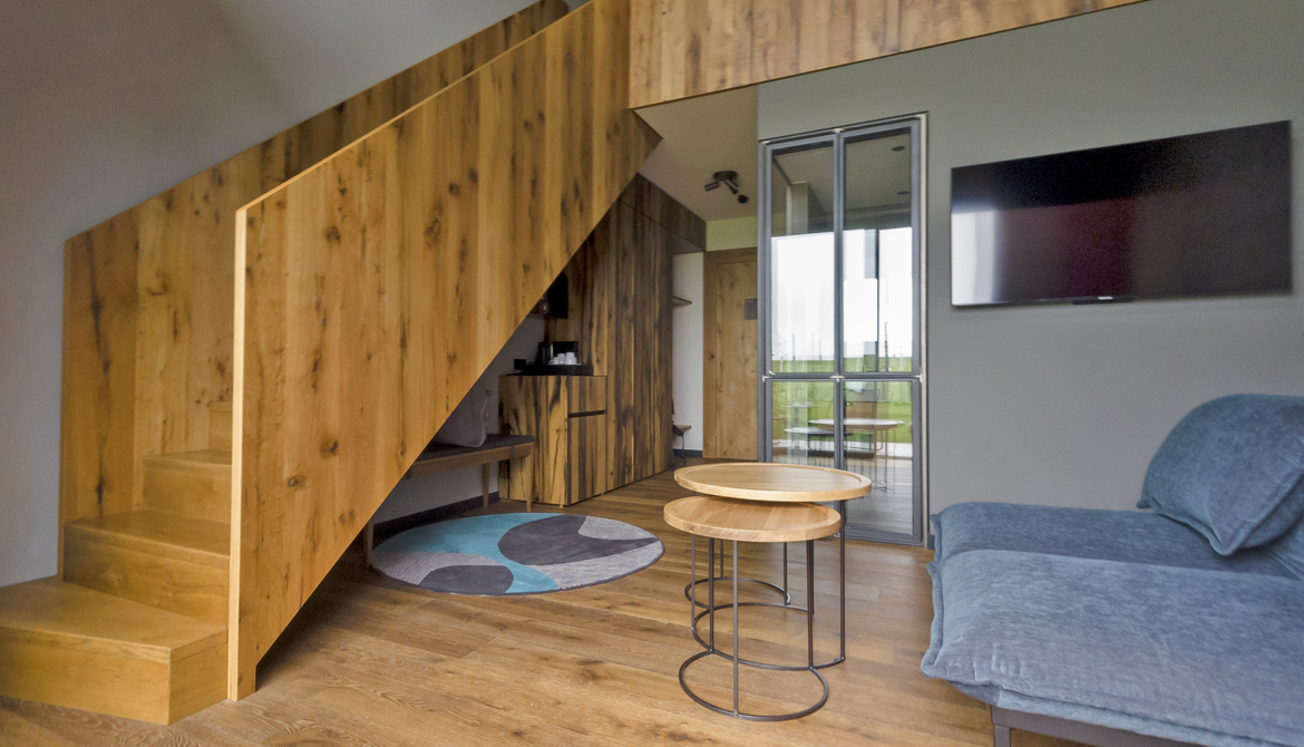 Modern living room with wooden staircase and floor, a blue sofa, two round, interlocking tables, a wall-mounted TV and a glass door leading onto a balcony. A colorful carpet accents the cozy furnishings.