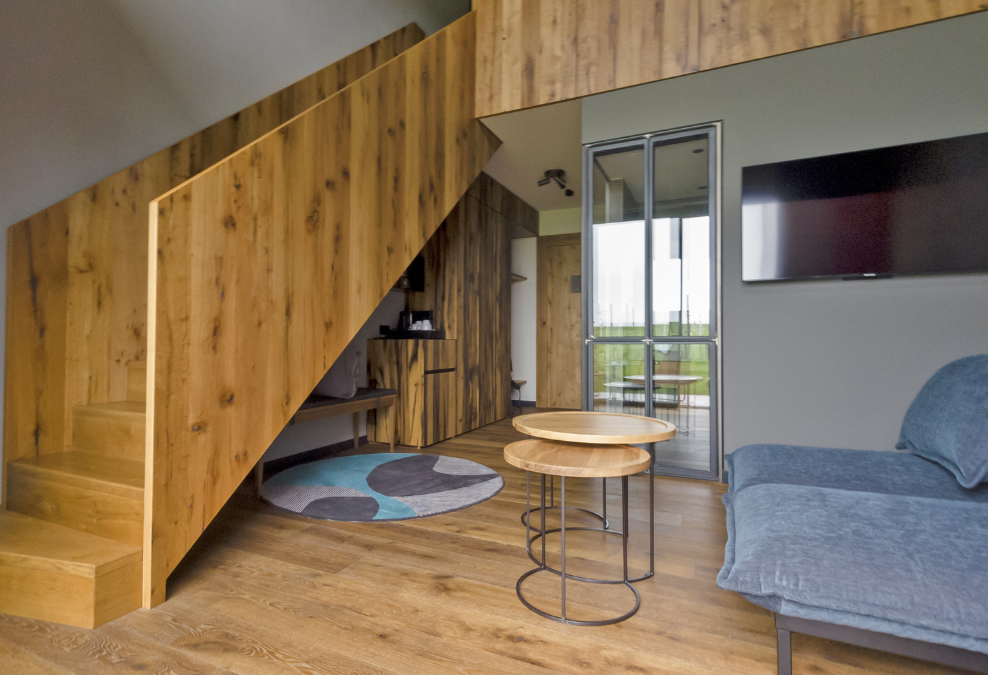 Modern living room with wooden staircase and floor, a blue sofa, two round, interlocking tables, a wall-mounted TV and a glass door leading onto a balcony. A colorful carpet accents the cozy furnishings.