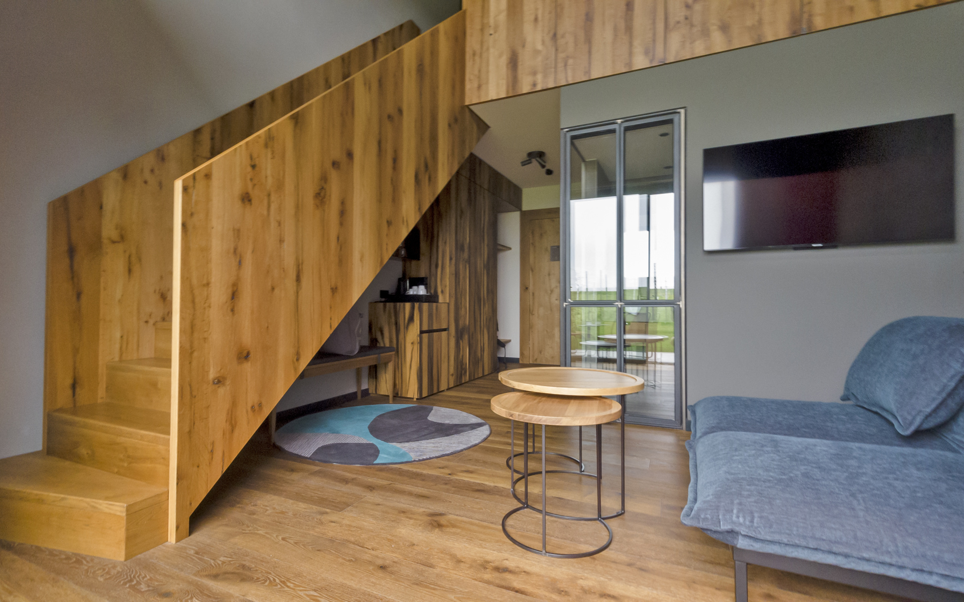 Modern living room with wooden staircase and floor, a blue sofa, two round, interlocking tables, a wall-mounted TV and a glass door leading onto a balcony. A colorful carpet accents the cozy furnishings.