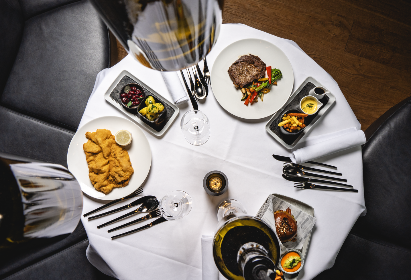 A table laid with steak, schnitzel, chips, vegetables and glasses of wine on a white tablecloth.