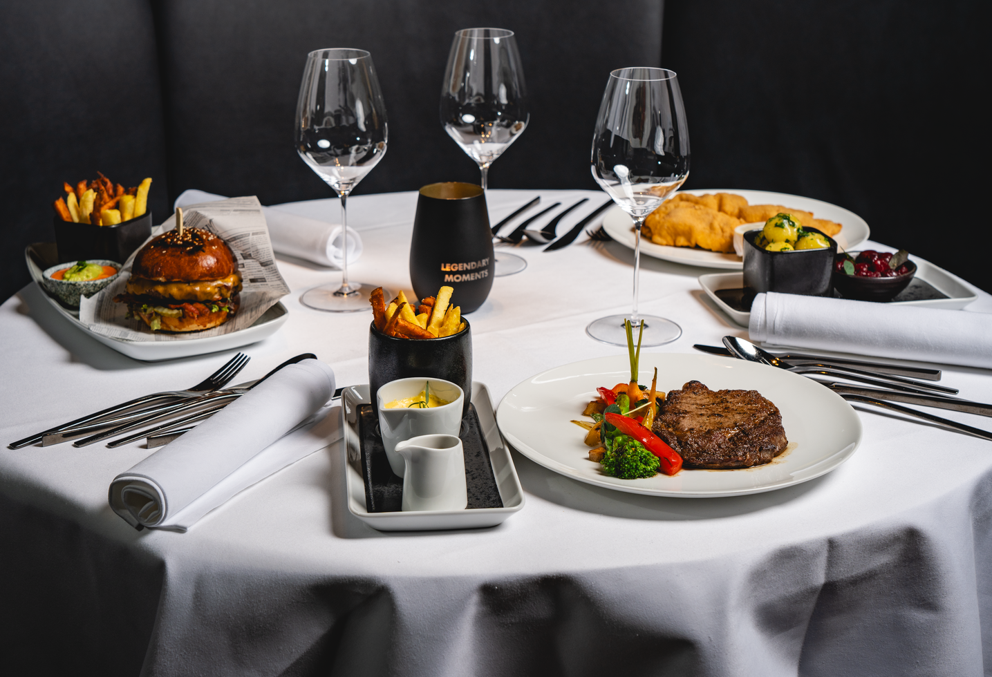 A round table with gourmet steak, burgers, chips, wine glasses and side dishes on a white tablecloth.