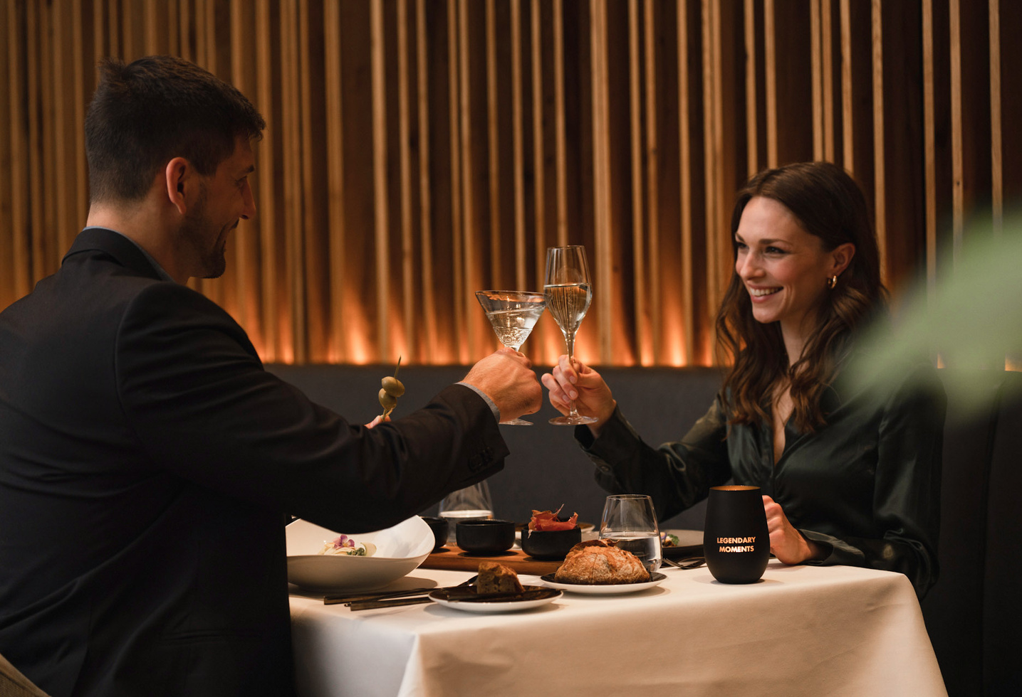 A smiling couple in formal dress clink glasses of wine at a candlelit restaurant table.