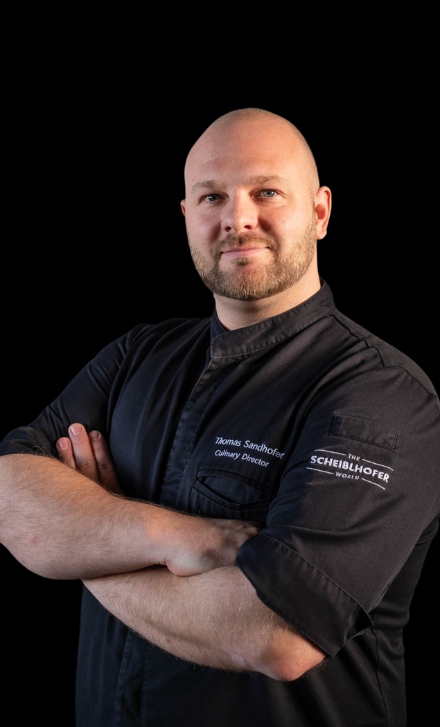 Bald man in a black chefs jacket with folded arms, looking confidently into the camera, in front of a black background.