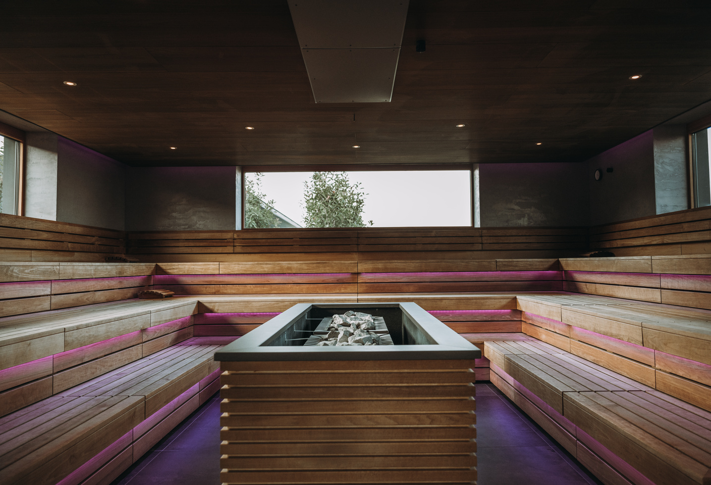 Moderne Holzsauna mit abgestuften Bänken, zentralem Steinofen und großen Fenstern, die natürliches Licht hereinlassen.