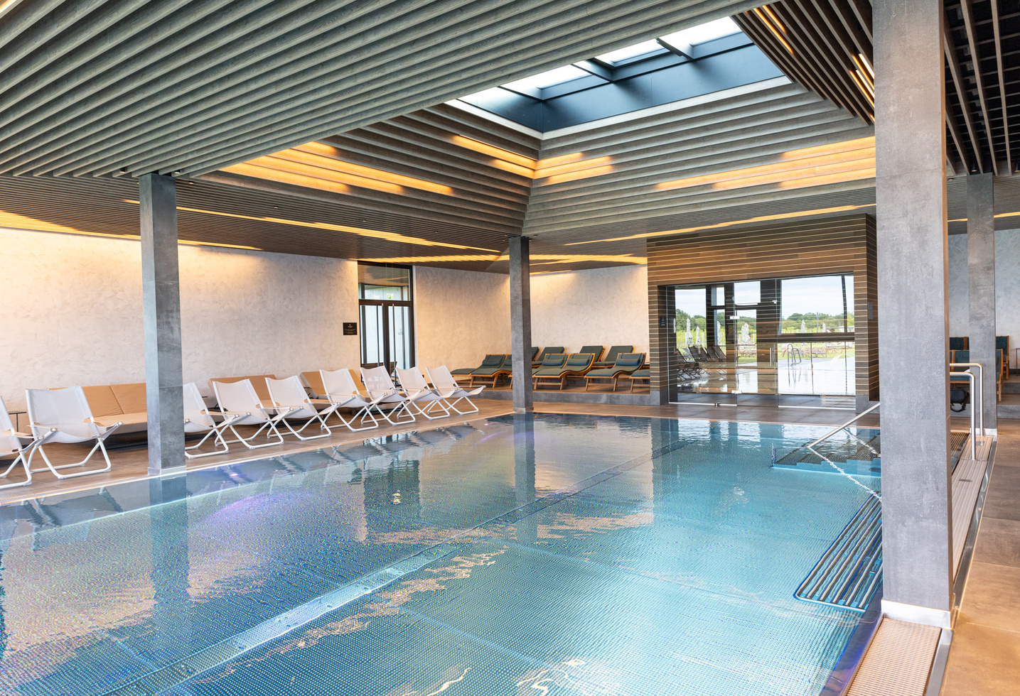 Hallenbad mit Liegestühlen, modernen Decken, großen Fenstern und natürlichem Licht, das in den Raum strömt.