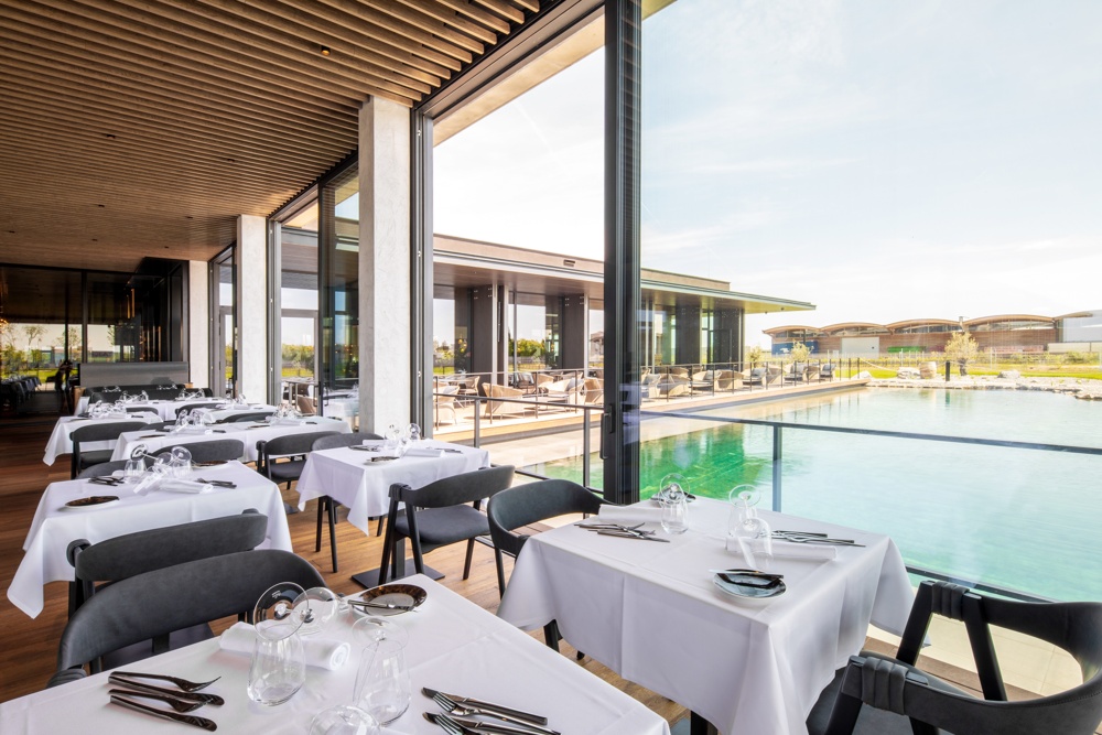 Modern restaurant with white tablecloths and large windows overlooking the pool and outdoor terrace.