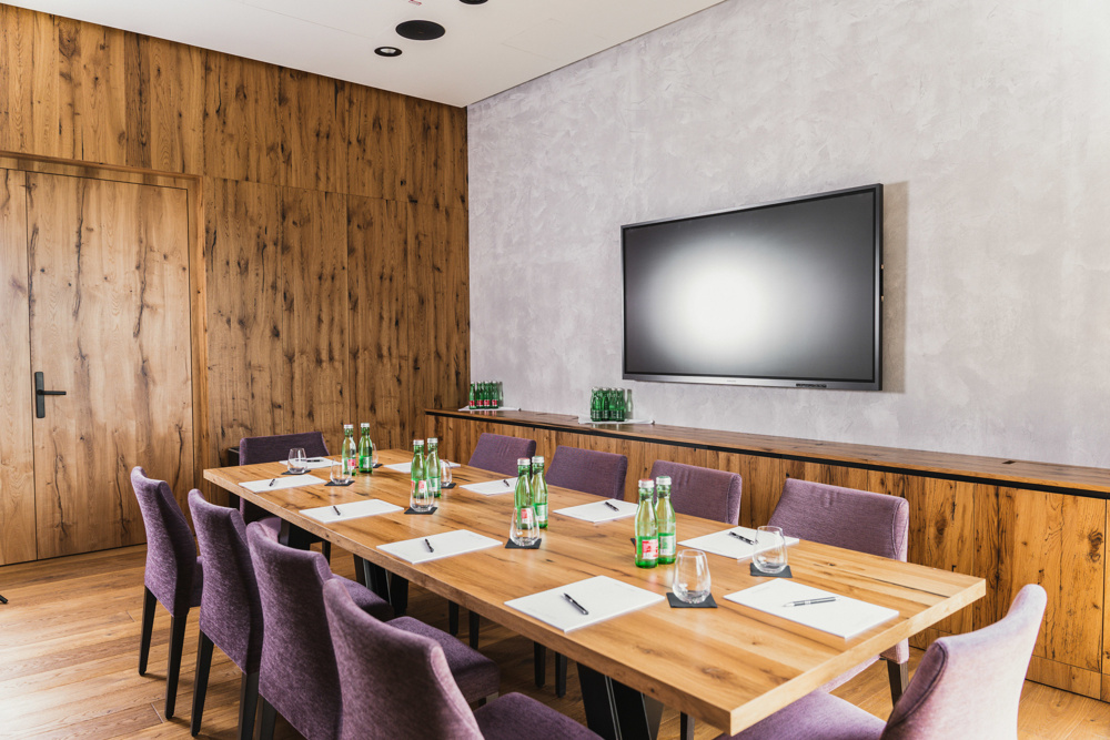 Moderner Tagungsraum mit Holztisch, lilafarbenen Stühlen, Wasserflaschen, Notizblöcken und einem an der Wand montierten Fernseher.