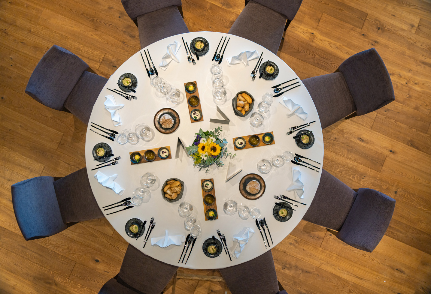 Ein runder, für acht Personen gedeckter Tisch mit Tellern, Gläsern, Servietten und einem Sonnenblumen-Mittelstück, von oben gesehen.