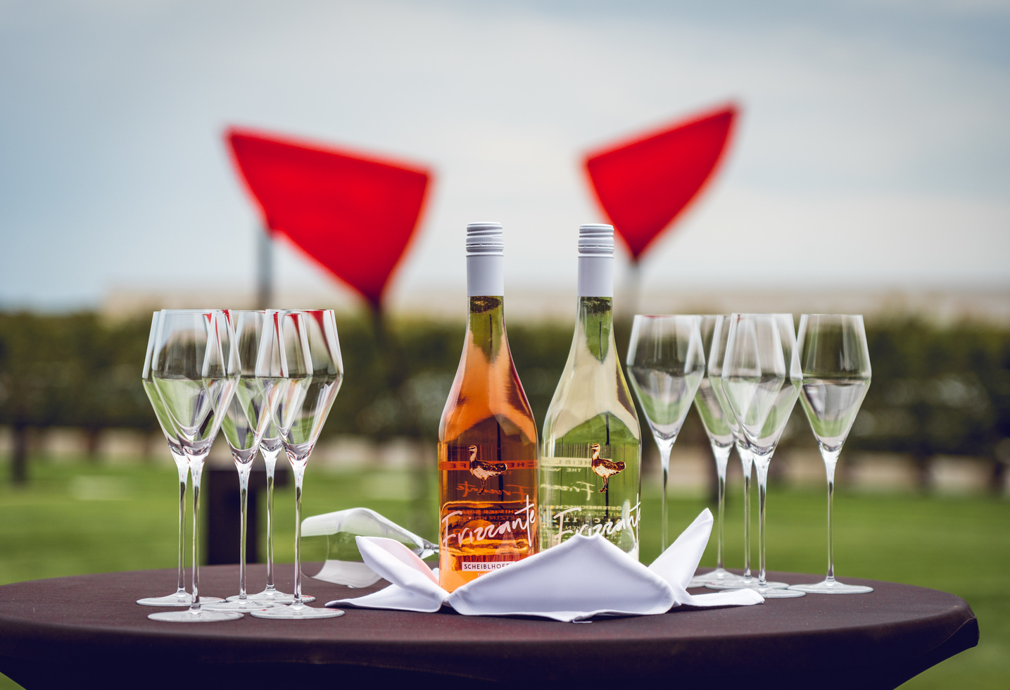 Zwei Weinflaschen und sechs leere Gläser auf einem Tisch im Freien mit roten Skulpturen im Hintergrund.
