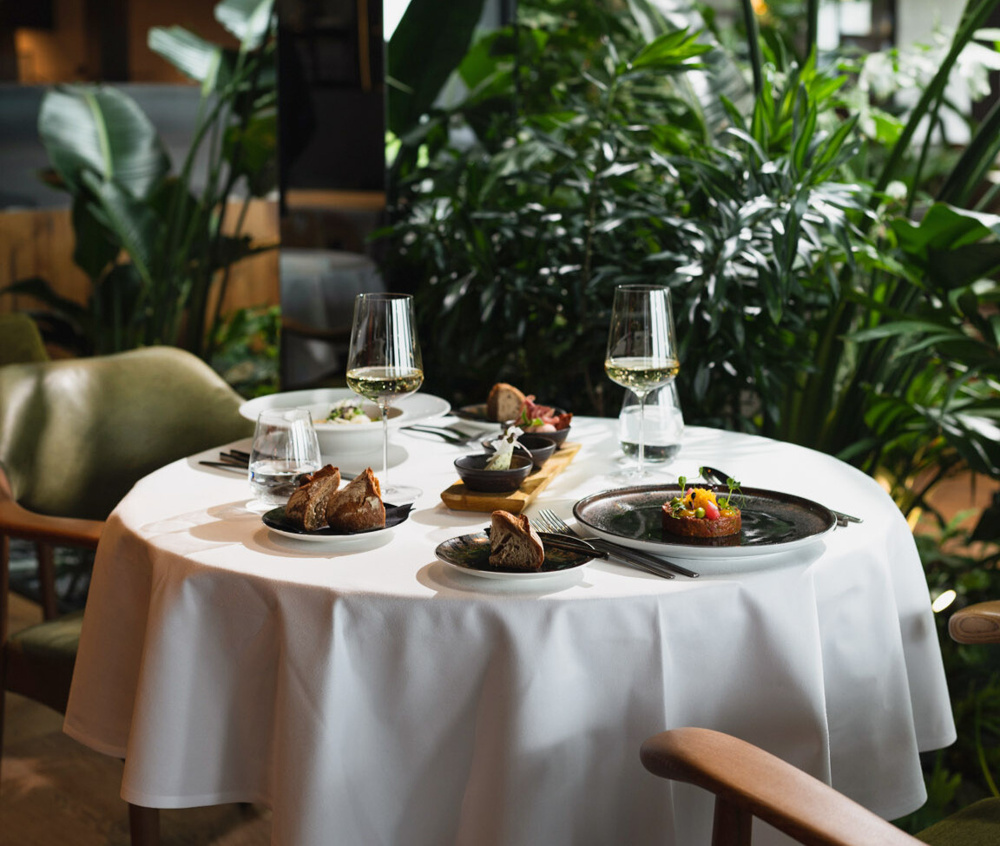Ein runder Tisch für zwei Personen mit Gourmetgeschirr und Weingläsern, umgeben von üppigen Grünpflanzen.