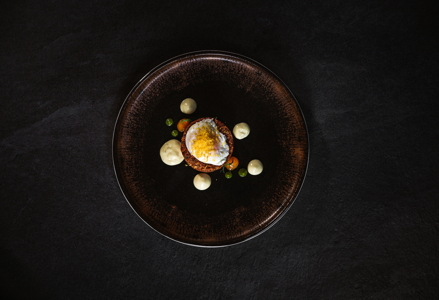 A gourmet dish with a poached egg and garnishes, artfully arranged on a dark round plate against a black background.