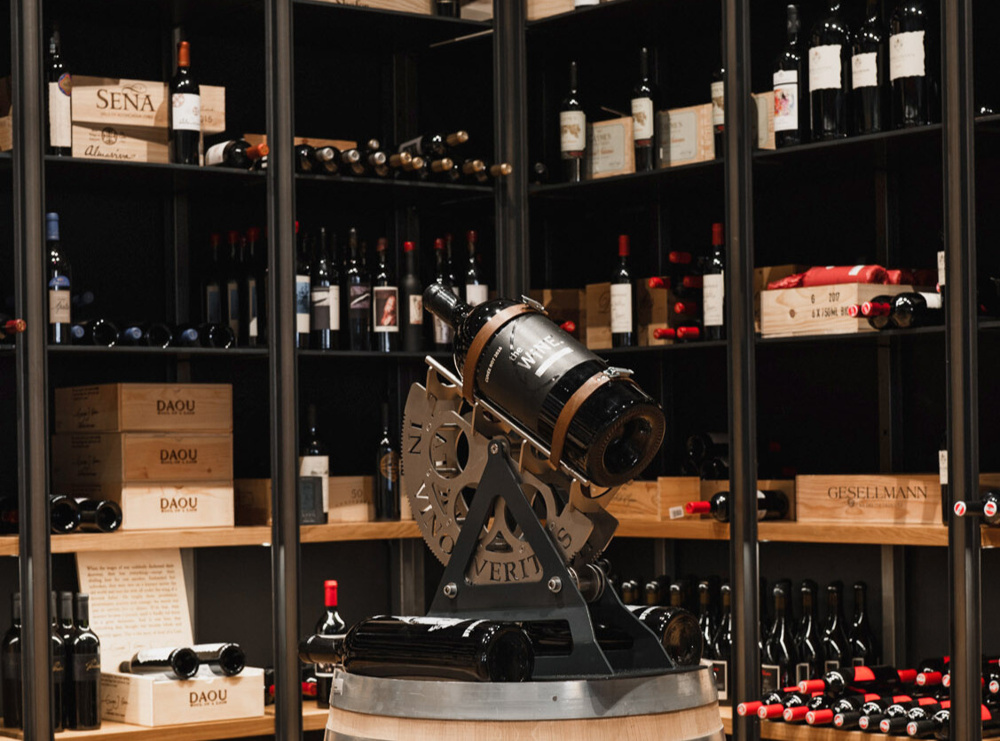 Große Weinflasche auf einem Metallständer vor Regalen, die mit verschiedenen Weinflaschen und Holzkisten gefüllt sind.