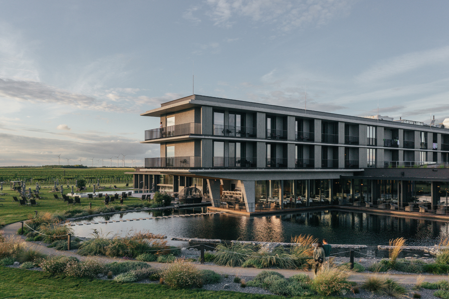 Modern three-story building with balconies, surrounded by water and greenery, under a partly cloudy sky.