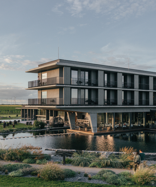 Modern three-story building with balconies, surrounded by water and greenery, under a partly cloudy sky.