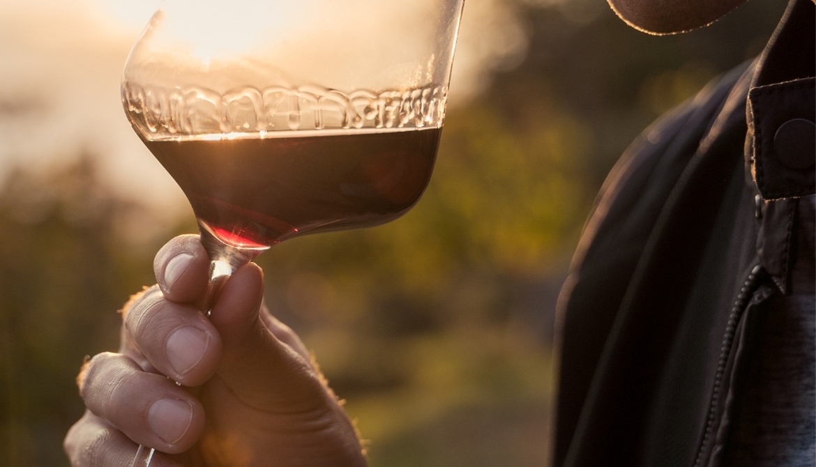 Person smelling a glass of red wine outdoors at sunset and holding the glass to their nose.