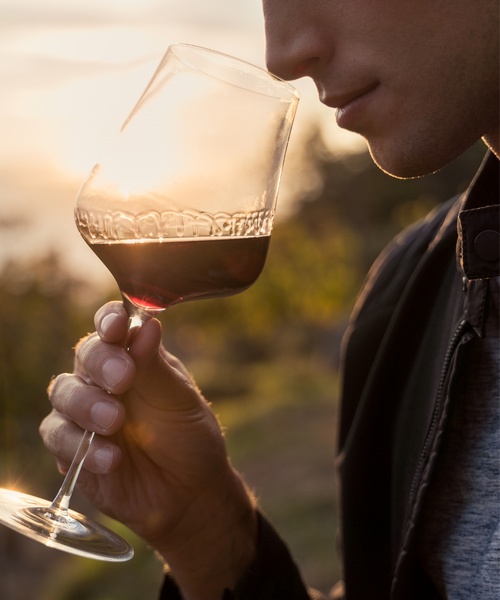 Person smelling a glass of red wine outdoors at sunset and holding the glass to their nose.